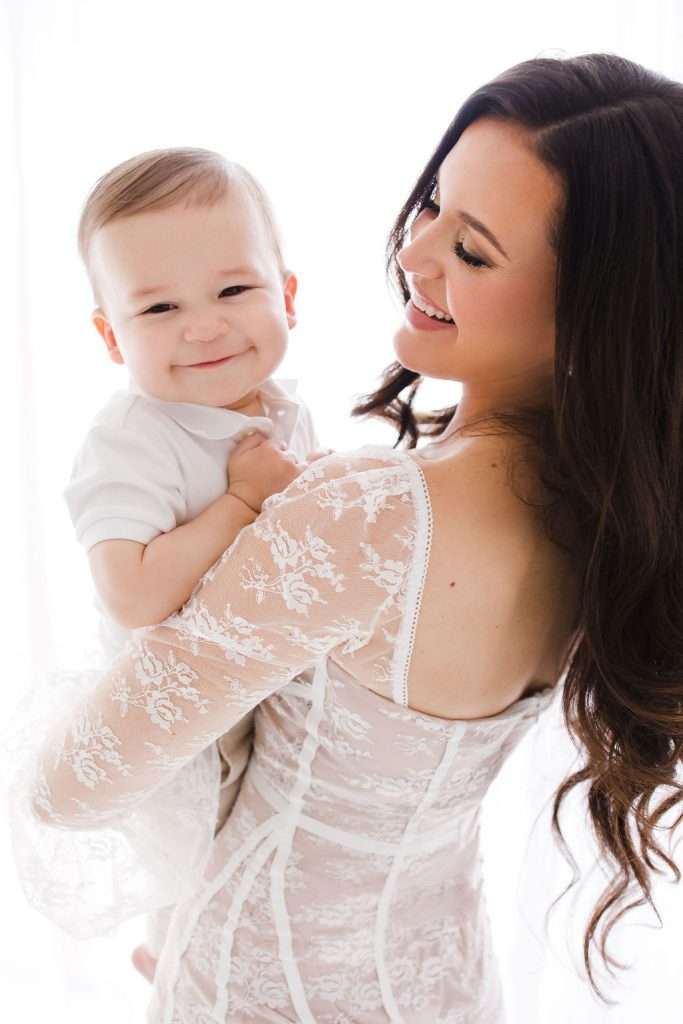 Woman in a white lace dress holding a smiling baby, both looking happy against a bright, white background.