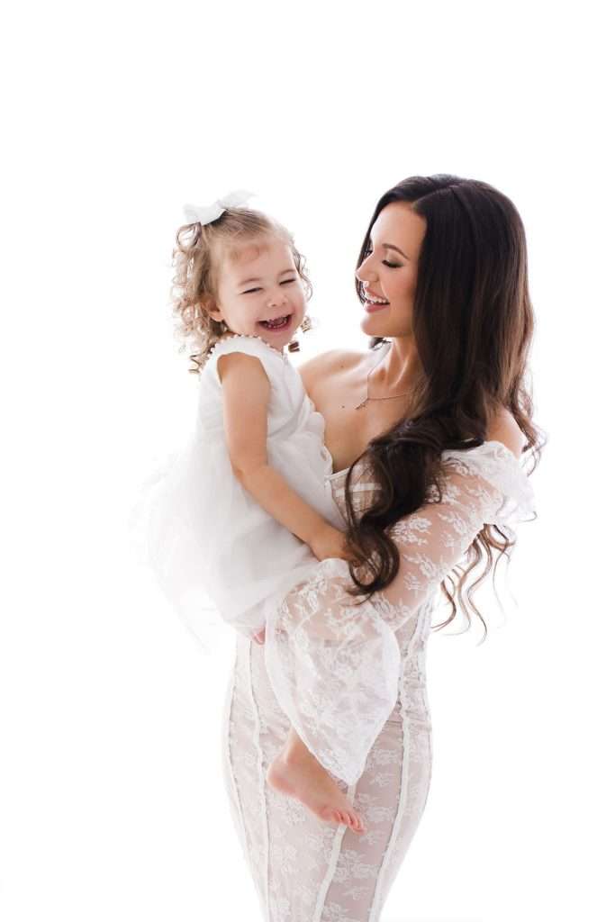 Woman in a white lace dress holds a smiling young girl in a white dress with a bow, both standing against a white background.