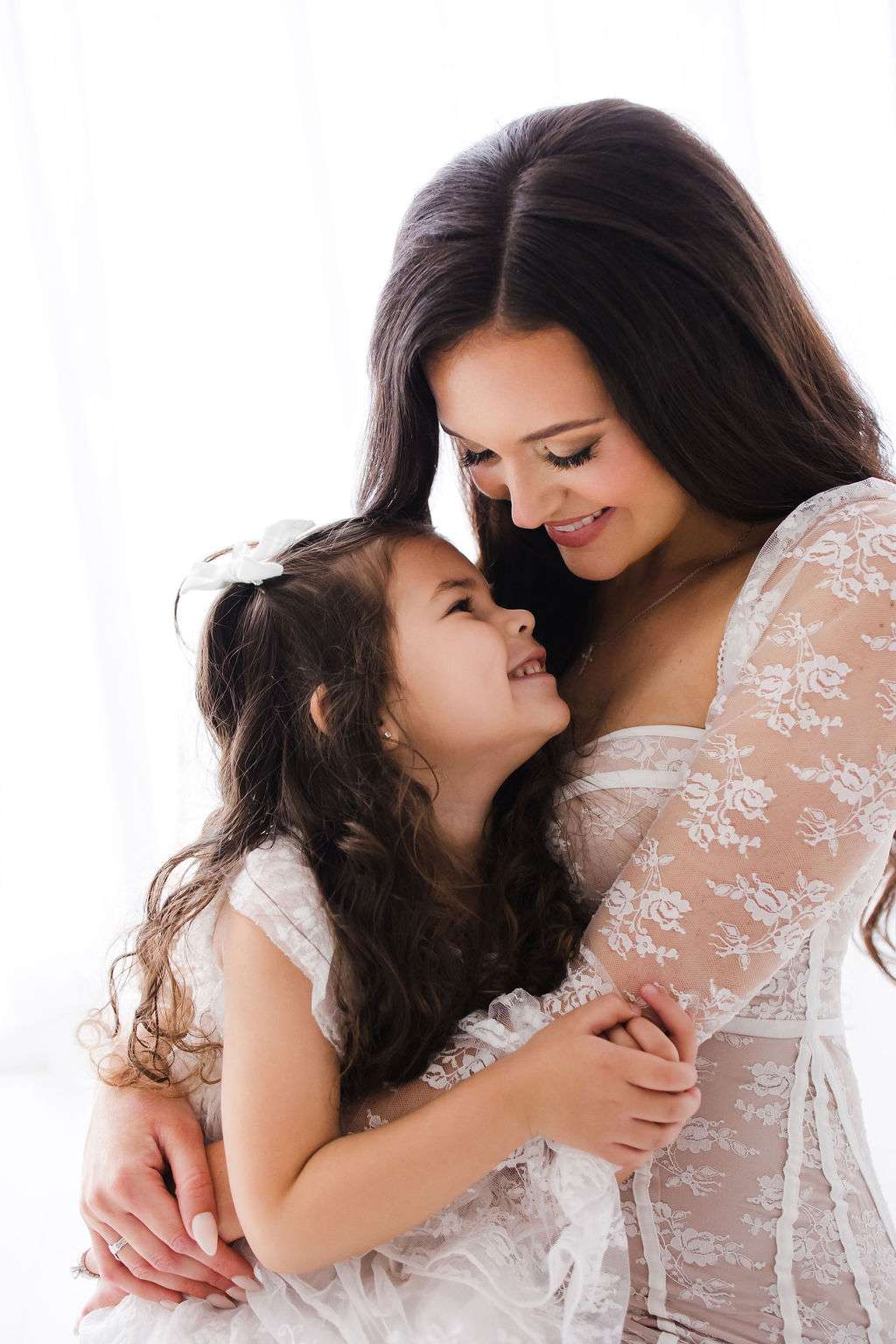 A woman in a lace dress hugs a smiling young girl with long hair; both are wearing white and appear happy in a brightly lit setting.