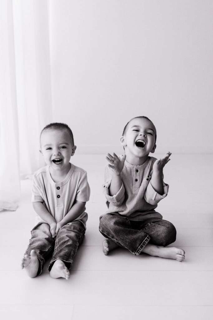 Two young boys sit barefoot on the floor, smiling and laughing. They wear casual jeans and long-sleeve shirts in a bright, minimal room.