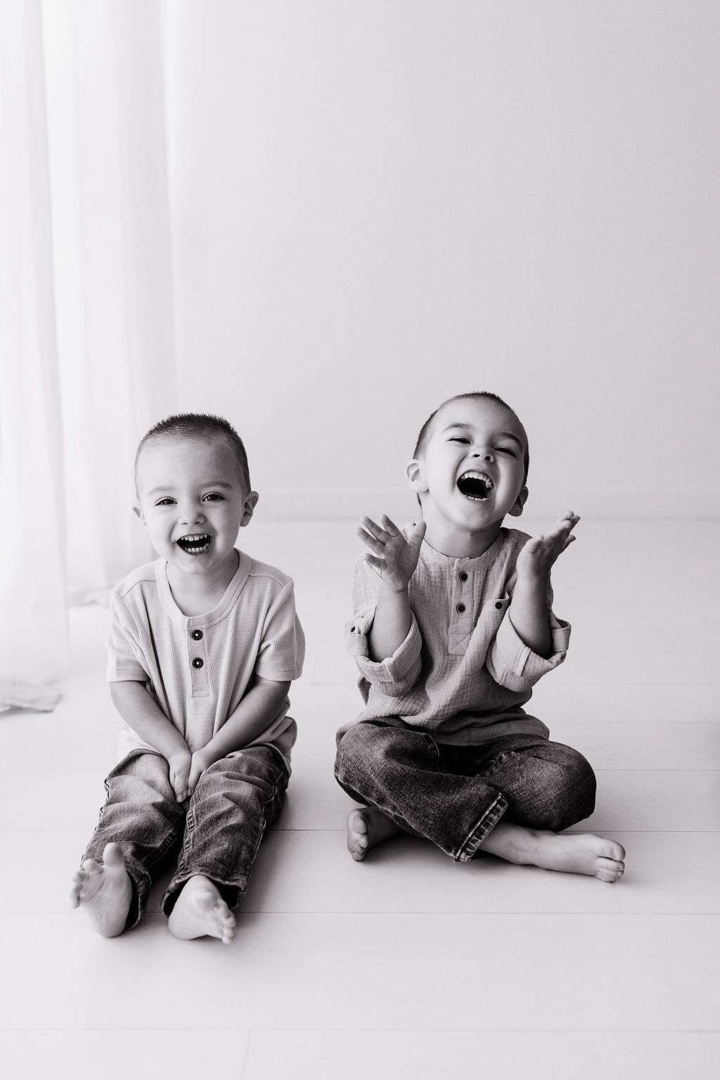 Two young boys sit barefoot on the floor, smiling and laughing. They wear casual jeans and long-sleeve shirts in a bright, minimal room.