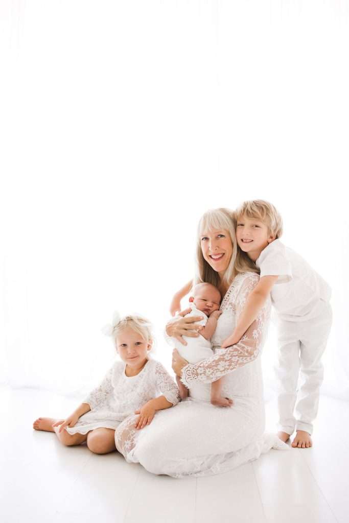 Woman in a white dress sits with three children, including a baby in her arms, a girl seated beside her, and a boy standing next to her. All are dressed in white, against a bright background.