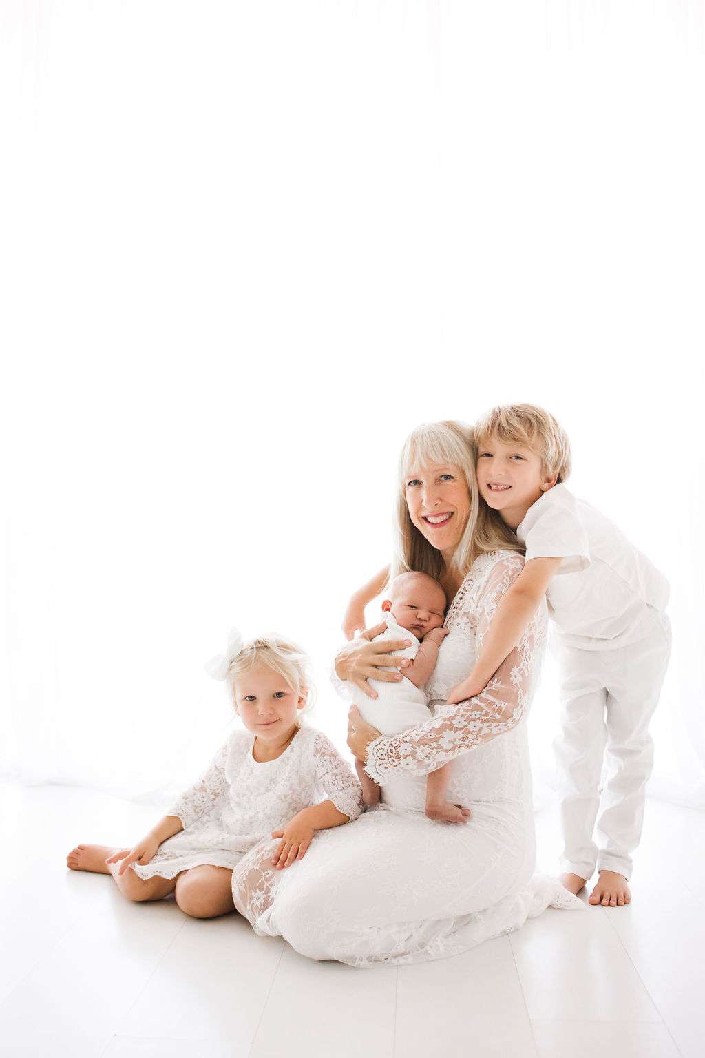 Woman in a white dress sits with three children, including a baby in her arms, a girl seated beside her, and a boy standing next to her. All are dressed in white, against a bright background.