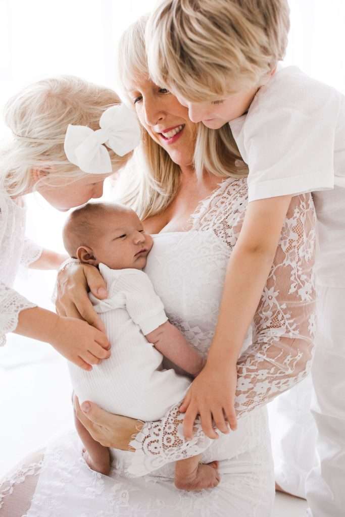 Woman in white lace dress holds a baby while two young children, one with a bow and another leaning in, embrace her and the baby. All are dressed in white, with a bright background.