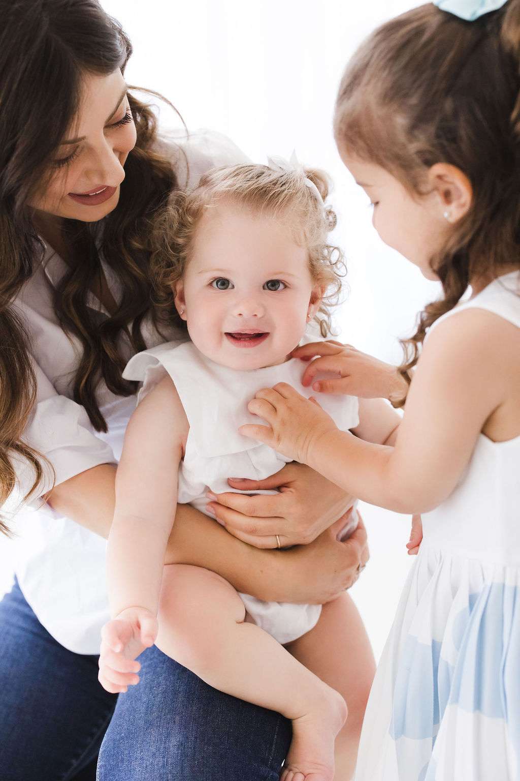 A woman holds a smiling baby while an older child, both girls wearing white, gently touches the baby’s chest. All appear happy in a bright setting.