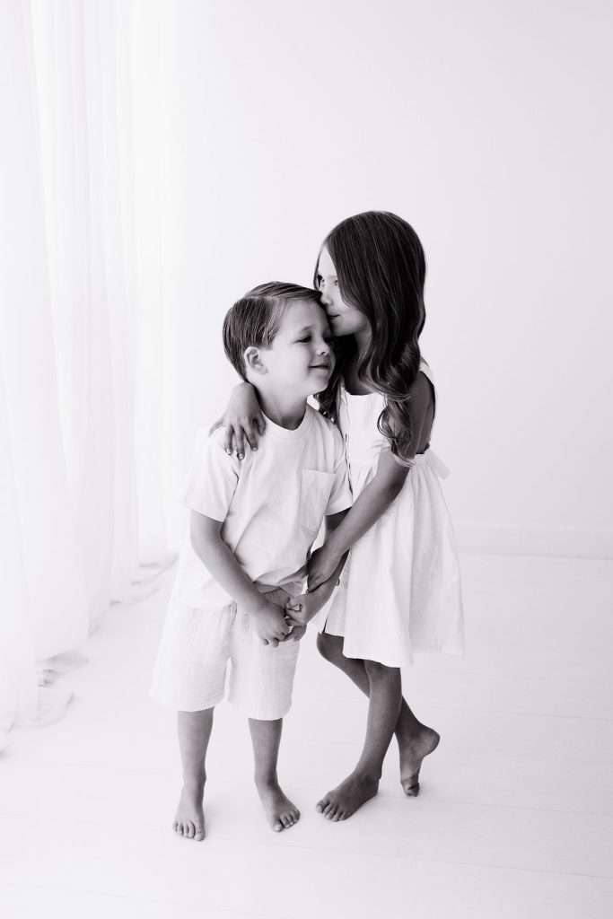 Young girl in a dress hugs and kisses a boy on the forehead. Both children are standing barefoot in a bright, white room.