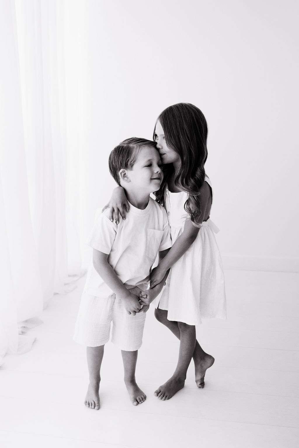 Young girl in a dress hugs and kisses a boy on the forehead. Both children are standing barefoot in a bright, white room.