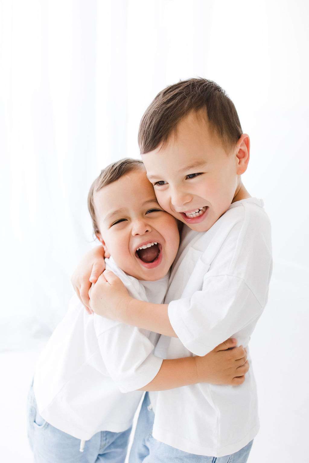 Two young children in white shirts and light jeans hug and smile against a bright, white background.
