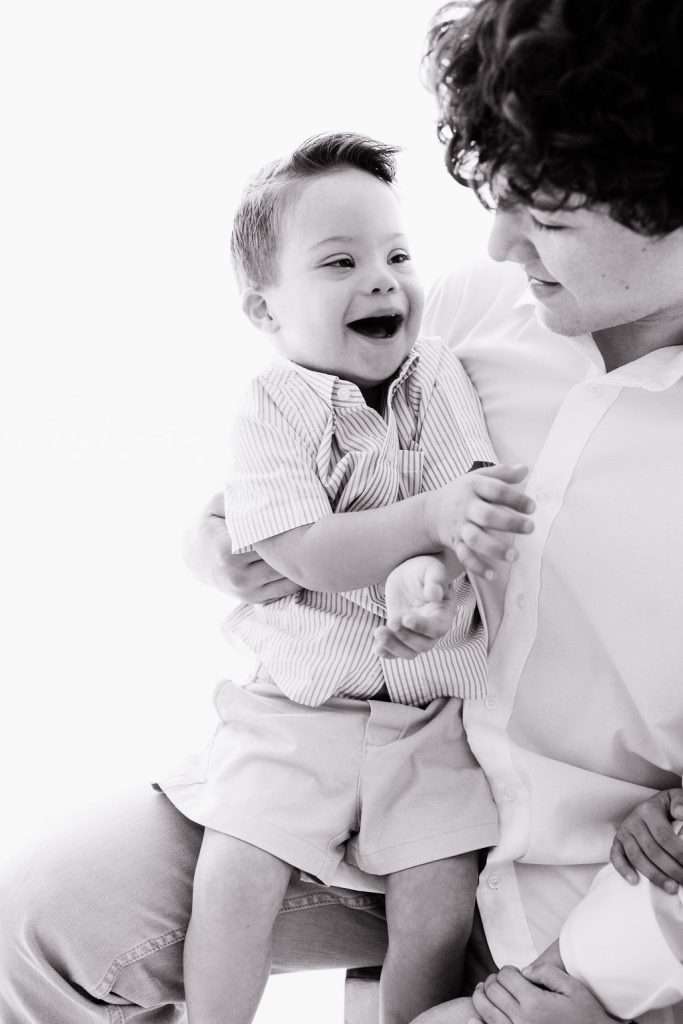 An adult holds a smiling young child on their lap. Both are dressed in light-colored clothing and appear to be interacting and making eye contact.