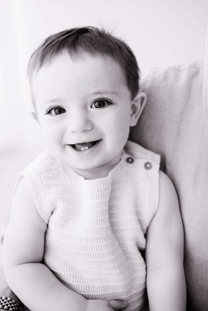 Black and white photo of a smiling baby with short hair, wearing a sleeveless textured top, sitting on a light-colored cushion.