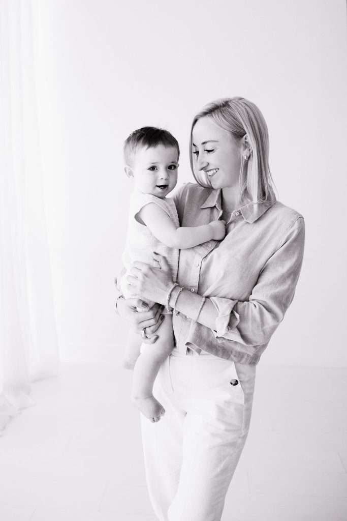 A woman smiles while holding a young child in her arms. Both are dressed in light-colored clothes in a bright, minimalistic room.