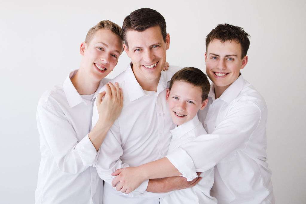 Four people, all wearing white shirts, are standing closely together and smiling at the camera against a plain white background.