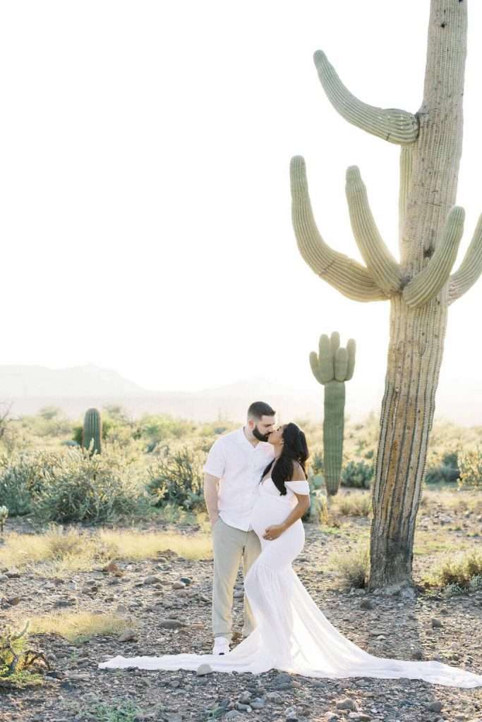 A couple stands in a desert landscape next to a tall cactus, with the woman in a white dress appearing pregnant as they kiss.