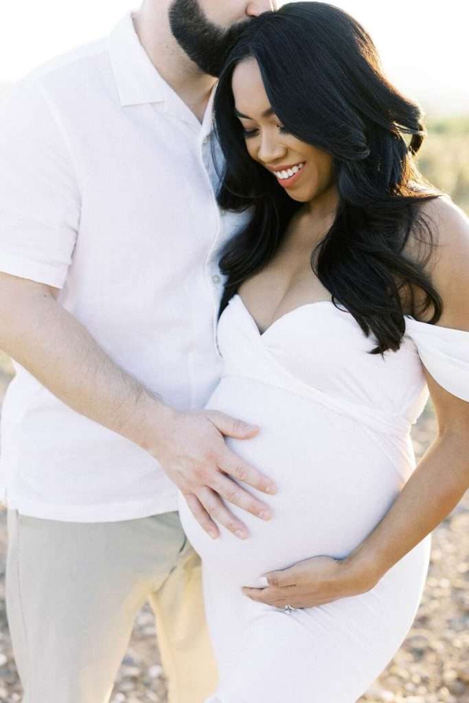 A man gently kisses a smiling pregnant woman’s head while placing his hand on her baby bump. Both are dressed in white and standing outdoors.