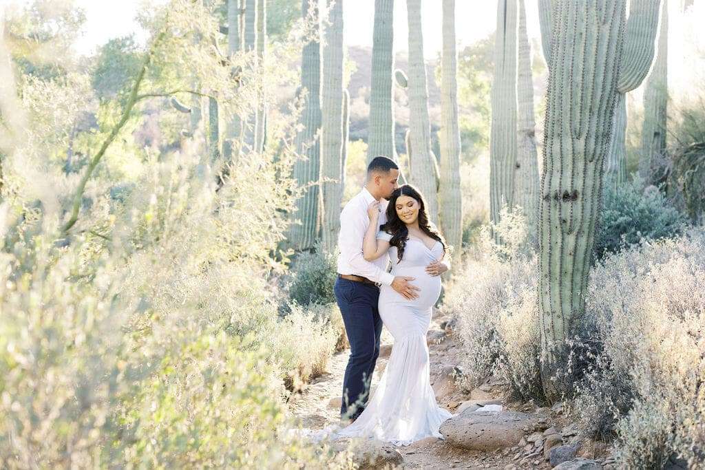 A couple stands among tall cacti in a desert landscape, with the man gently holding the pregnant woman's waist as she cradles her belly.