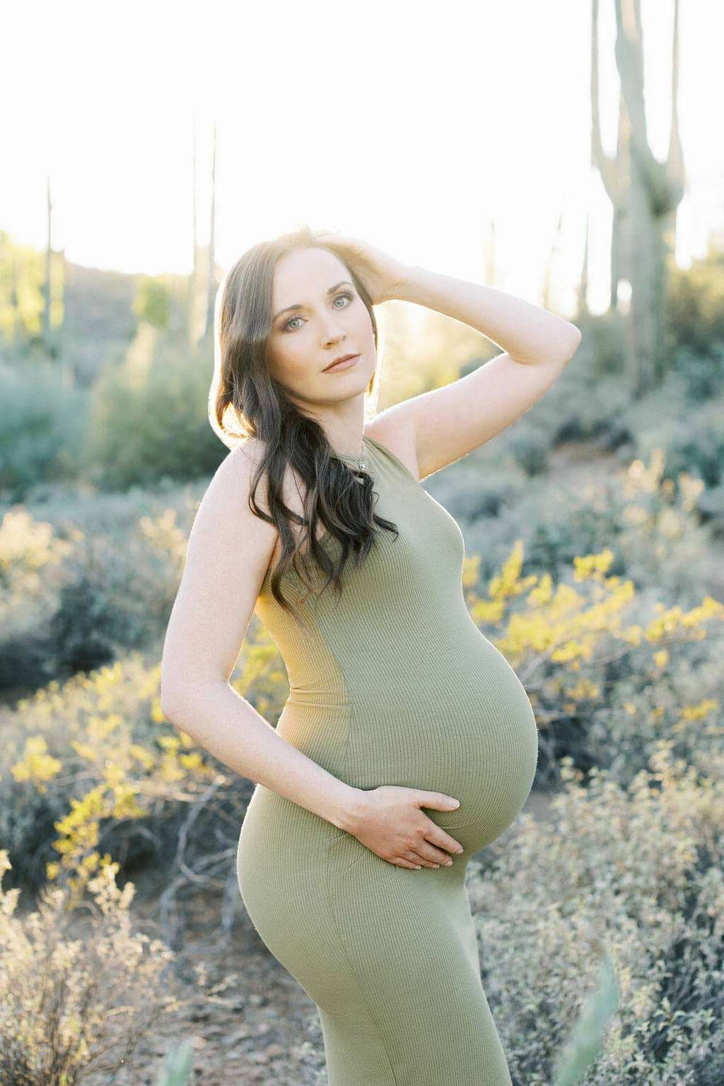 A pregnant woman in a green dress stands outdoors in a sunlit, desert-like landscape, holding her belly with one hand and touching her hair with the other.