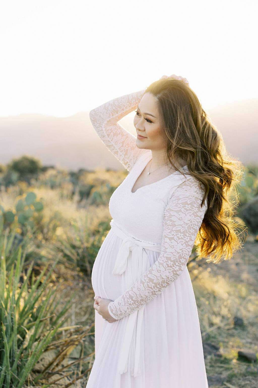 Pregnant woman in a white lace dress stands outdoors at sunset, holding her belly with one hand and touching her hair with the other, surrounded by desert plants.