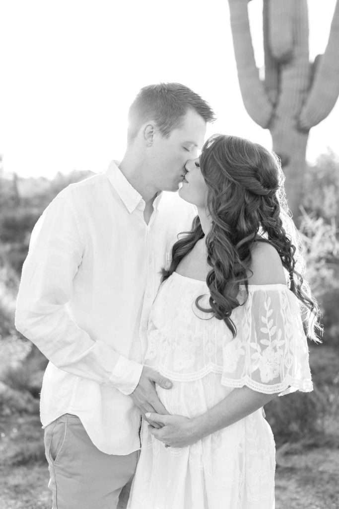 A couple stands outdoors, kissing, with the man touching the woman's pregnant belly. They both wear light-colored clothing, and a large cactus is visible in the background.