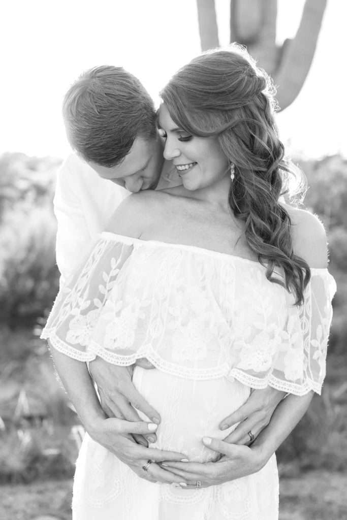 A couple stands close together outdoors, with the man embracing the pregnant woman from behind. Both have their hands on her belly and are smiling.