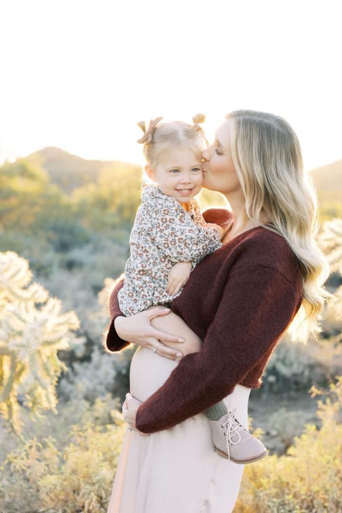 Woman holding a young girl outdoors, kissing her on the forehead. The girl is smiling and they are surrounded by sunlit plants and hills in the background.
