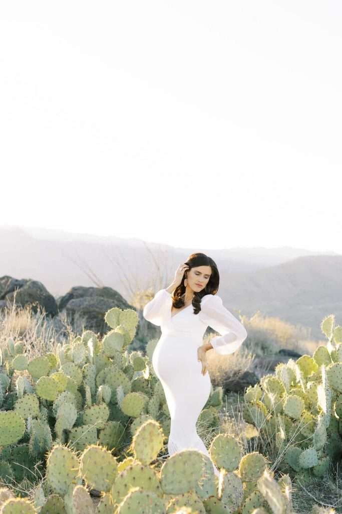Pregnant woman in a long white dress stands among cactus plants in a sunlit desert landscape with mountains in the background.