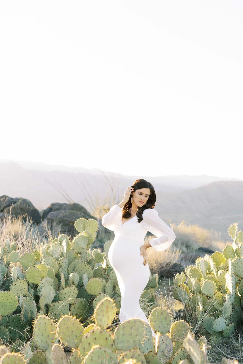 Pregnant woman in a long white dress stands among cactus plants in a sunlit desert landscape with mountains in the background.