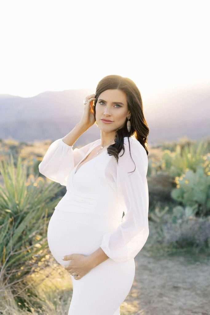 Pregnant woman in a white dress stands outdoors in a desert landscape at sunset, gently touching her belly and looking at the camera.
