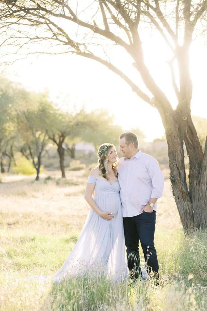 A pregnant woman in a light purple dress stands in a grassy field beside a man. They are looking at each other, with sunlight streaming through the trees behind them.