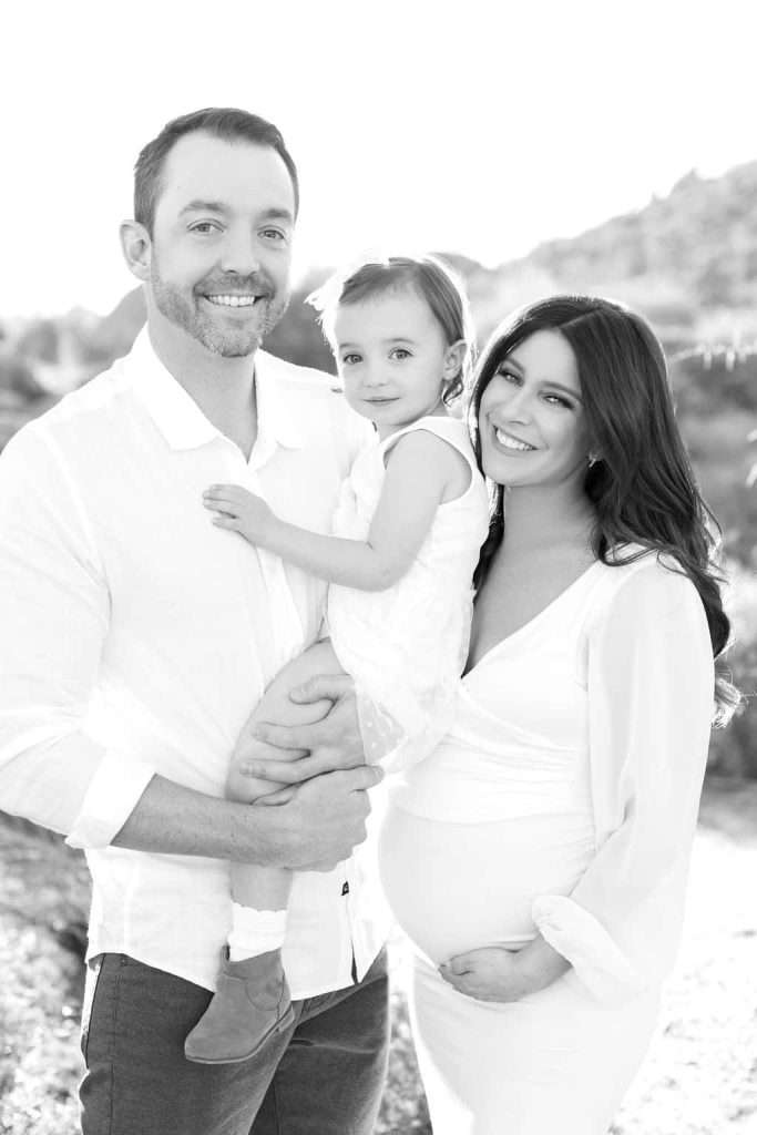 A man holds a young child while standing next to a pregnant woman outdoors; all three are dressed in light-colored clothing and smiling at the camera.