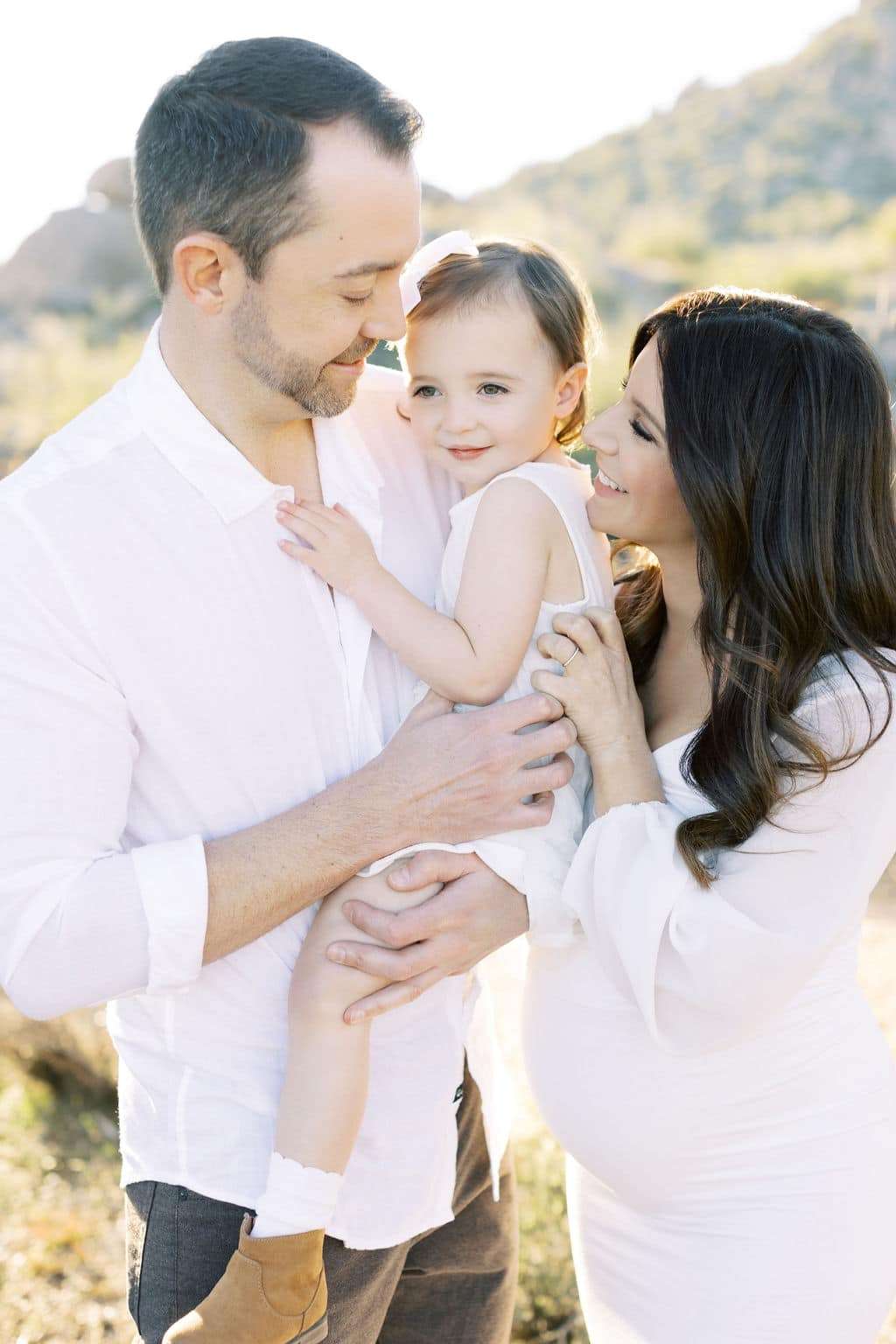 A man holds a young child while a pregnant woman stands beside them outdoors; all three are dressed in white and smiling.