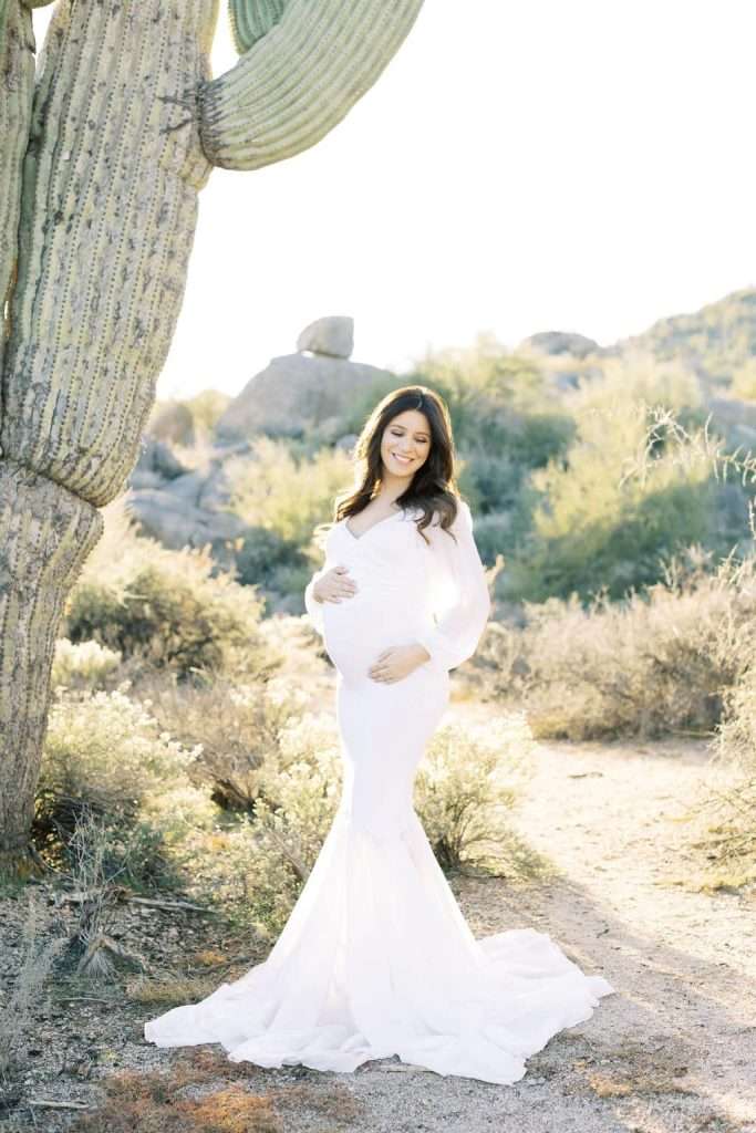 A pregnant woman in a long white dress stands outdoors in a desert landscape beside a large cactus, smiling and holding her belly.