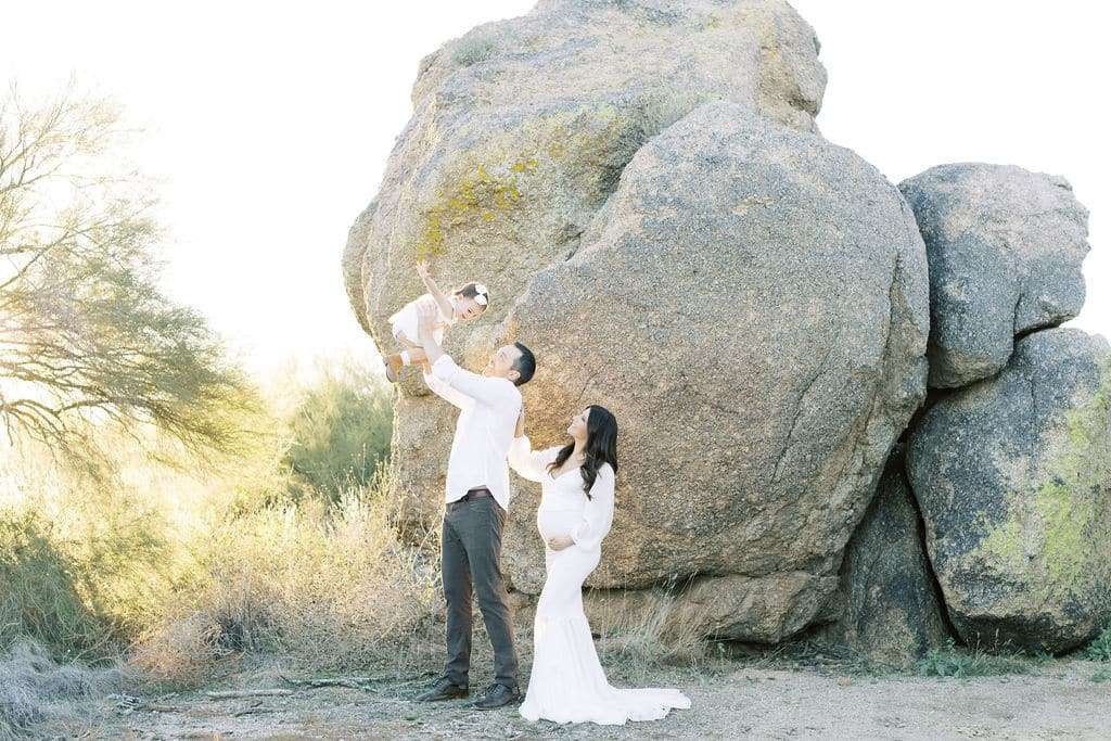 A man lifts a young child into the air while a woman stands beside them, all in front of large boulders in a sunlit outdoor setting.