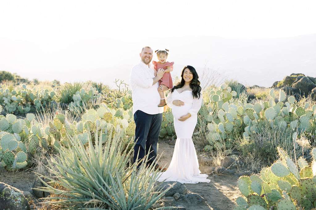 A man, woman, and young child stand together in a desert landscape with cactus plants. The woman is wearing a white dress and appears pregnant. The man is holding the child.