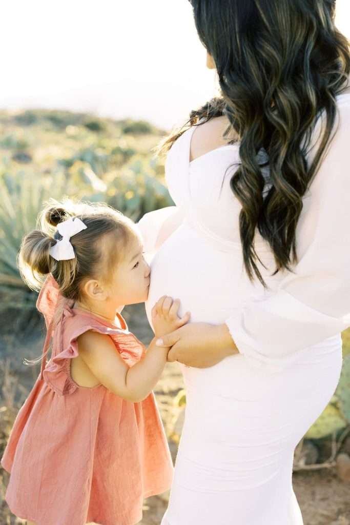 A young girl in a pink dress kisses the baby bump of a pregnant woman in a white dress outdoors.