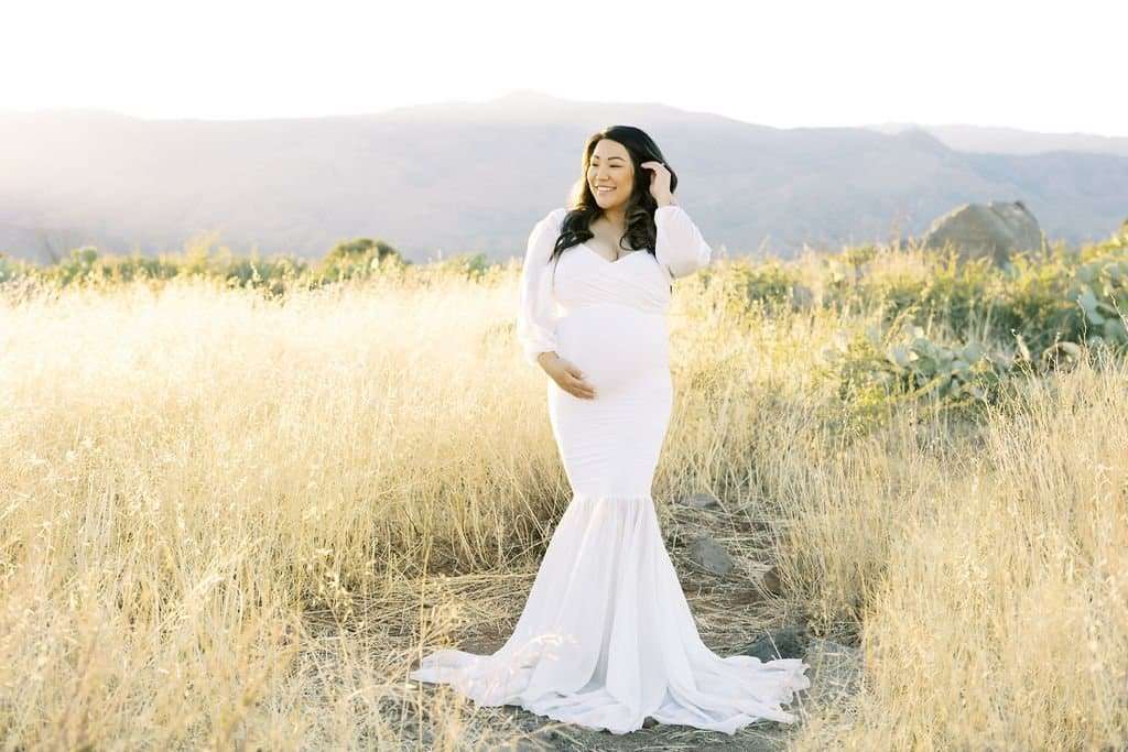 A pregnant woman in a long white dress stands in a sunlit field of tall grass with mountains in the background.