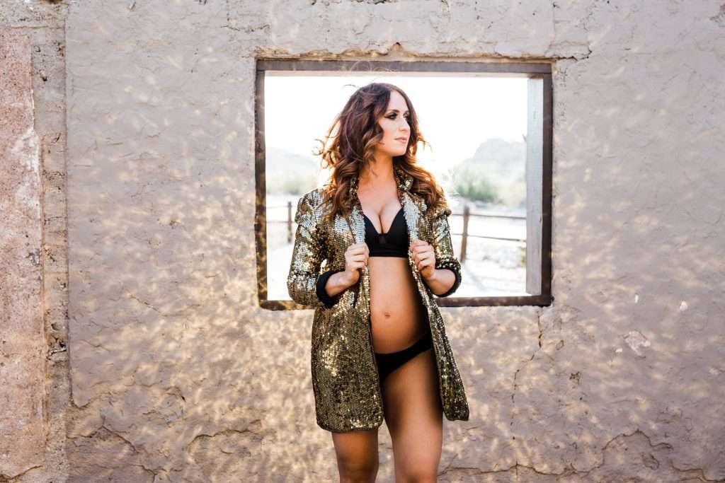 Pregnant woman in a black bikini and gold sequin jacket stands in a window frame of a weathered building, looking to the side.