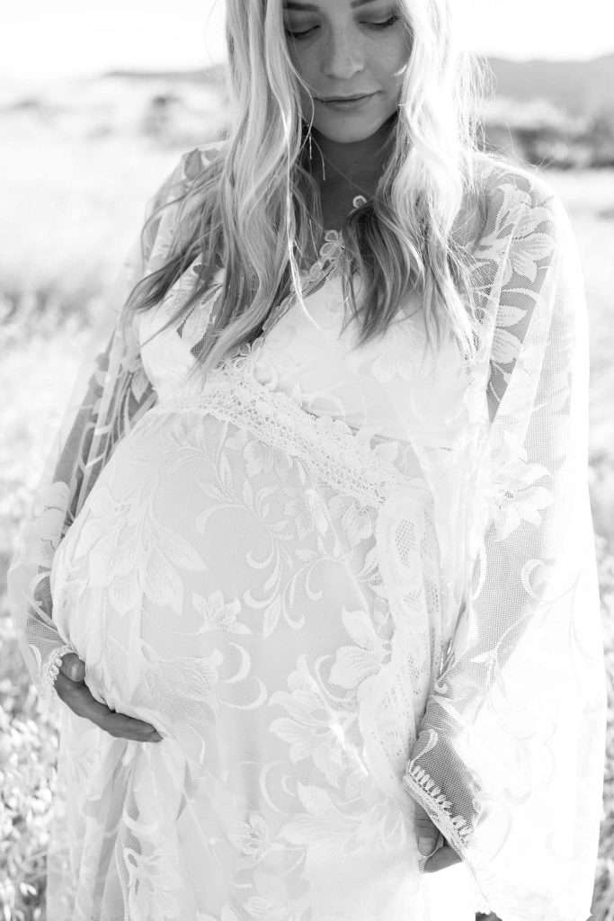 A pregnant woman in a lace dress stands outdoors, cradling her belly with both hands.