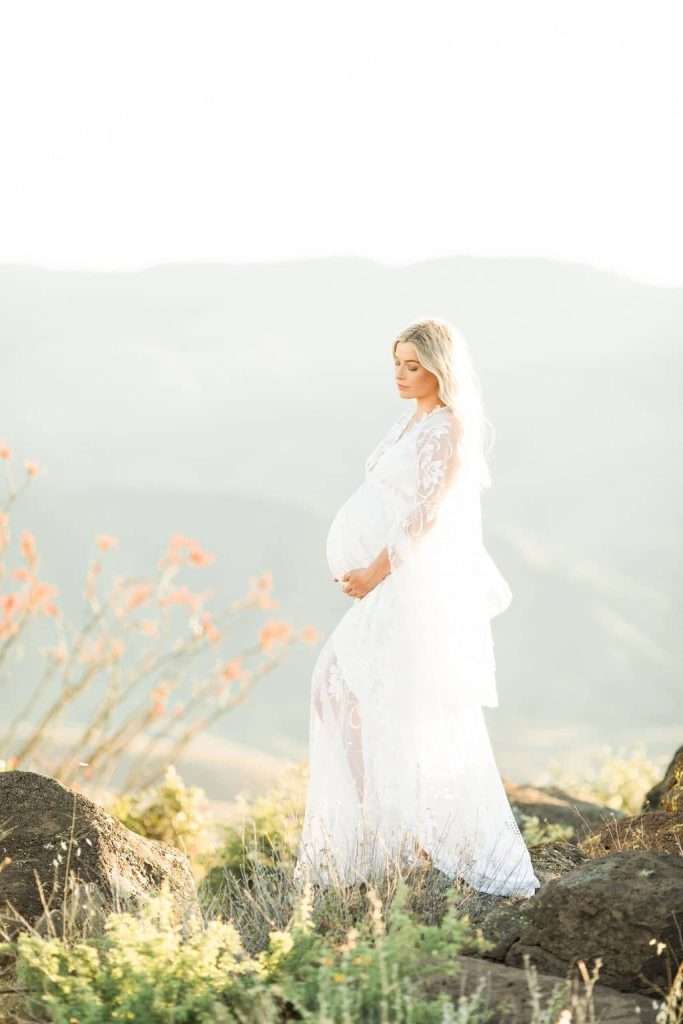 A pregnant woman in a white lace dress stands outdoors on rocky terrain, with mountains and wildflowers in the background.