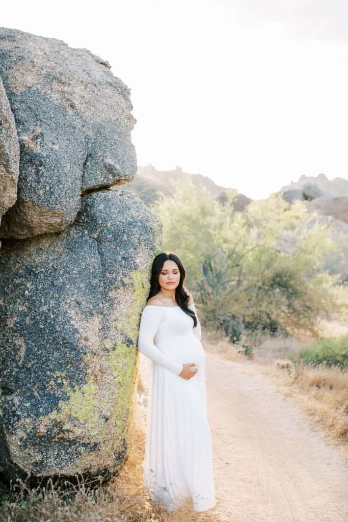 A pregnant woman in a long white dress stands outdoors, leaning against a large rock on a dirt path surrounded by nature.