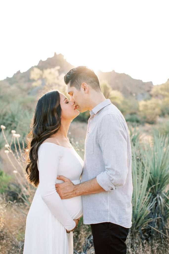 A couple stands in a natural outdoor setting, kissing, while the man gently holds the woman's pregnant belly.