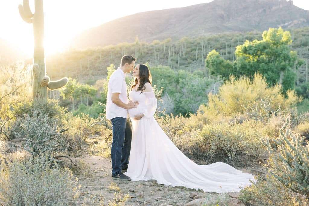 A couple stands in a desert landscape at sunset; the woman wears a long white dress and holds her pregnant belly while facing the man.