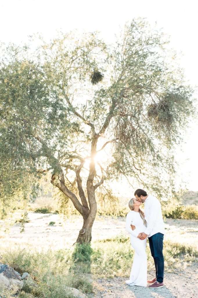 A couple stands closely together under a large tree, with sunlight shining through the branches in a natural outdoor setting.