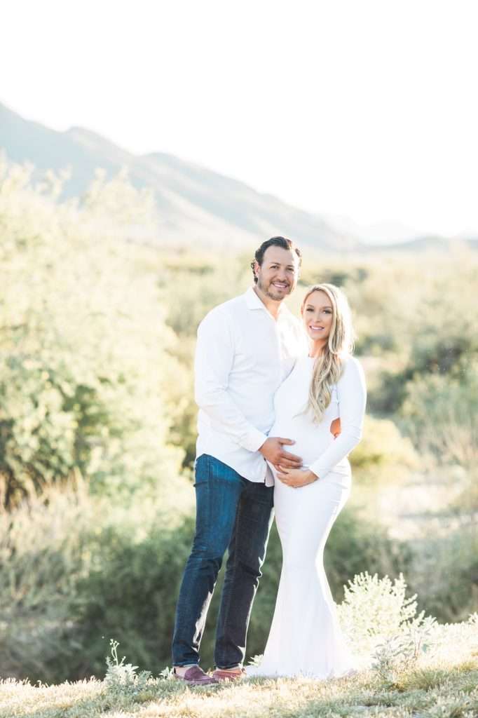 A man and a pregnant woman stand outdoors in a natural setting, both dressed in white, smiling and posing together.