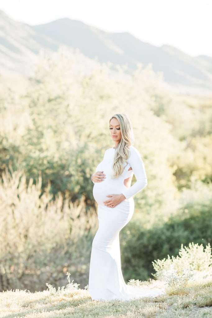 A pregnant woman in a long white dress stands outdoors in front of greenery and mountains, cradling her belly with both hands.