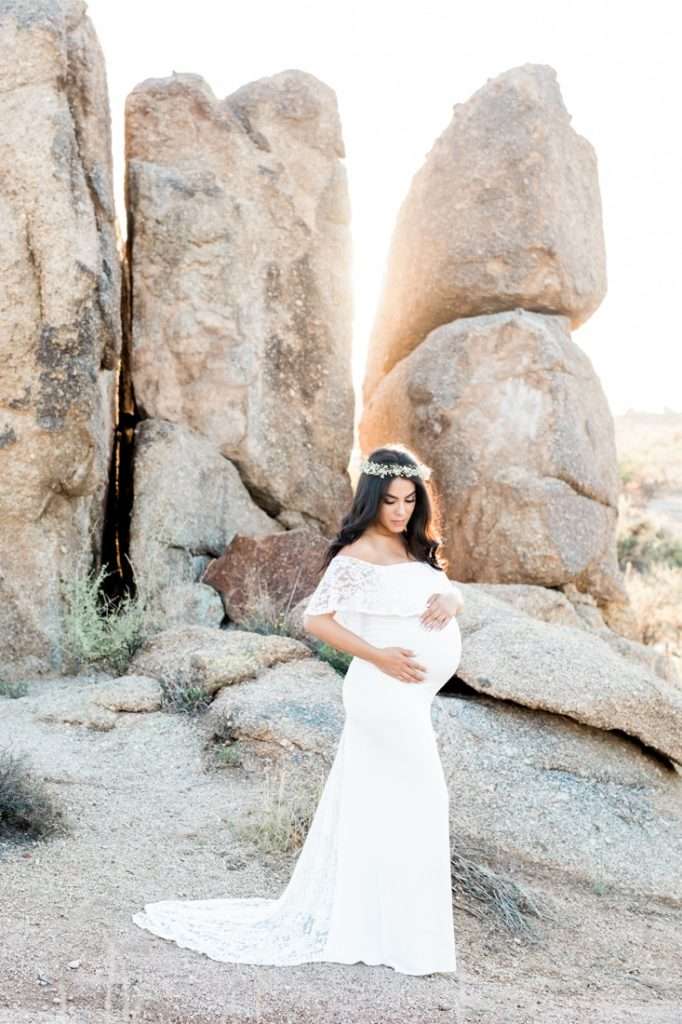 A pregnant woman in a white dress and floral crown stands in front of large rock formations outdoors, holding her belly.