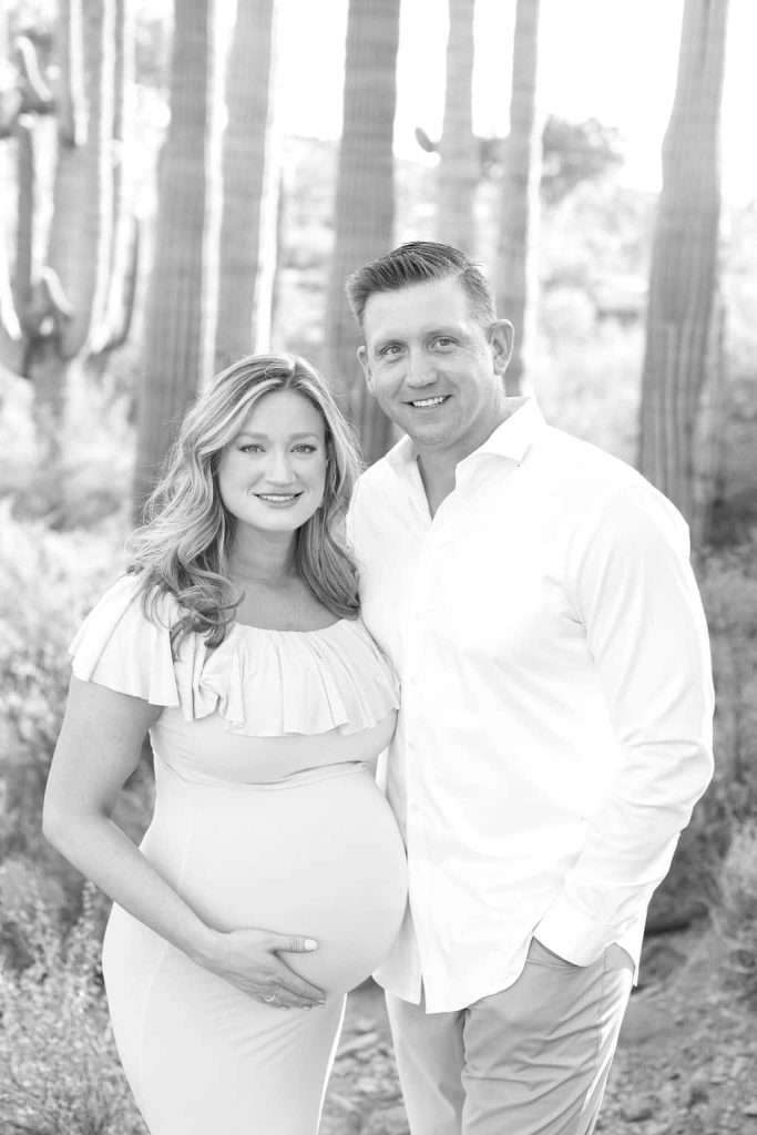 A pregnant woman and a man stand together outdoors in front of tall cacti, both smiling at the camera.