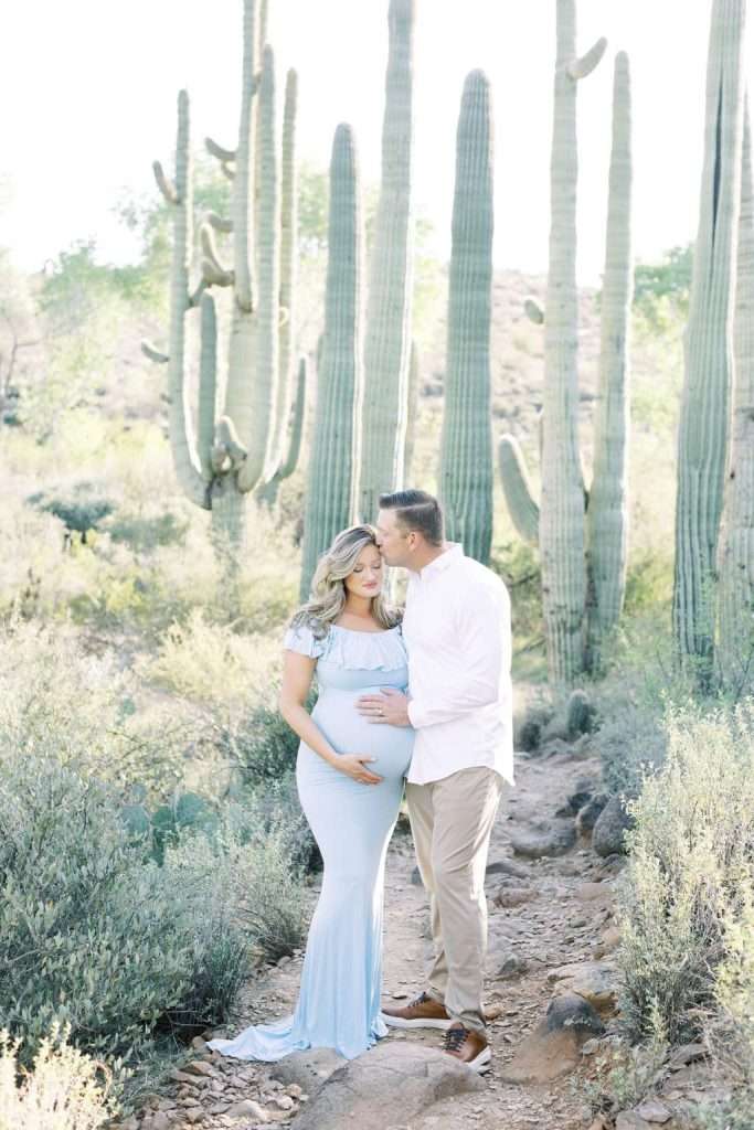 A pregnant woman in a light blue dress stands beside a man in a white shirt and khakis on a desert path with tall cacti in the background.
