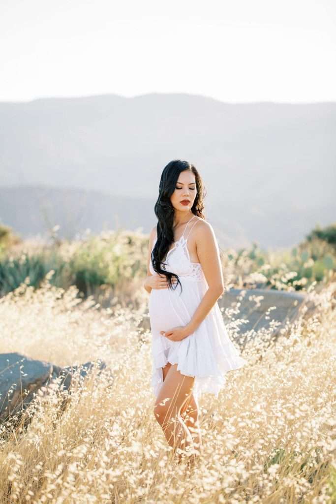 A pregnant woman in a white dress stands in a sunlit field of tall grass, with mountains visible in the background.