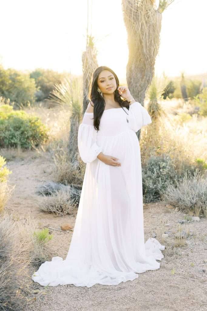 A woman in a long, flowing white dress stands outdoors in a desert landscape, gently holding her pregnant belly and looking at the camera.