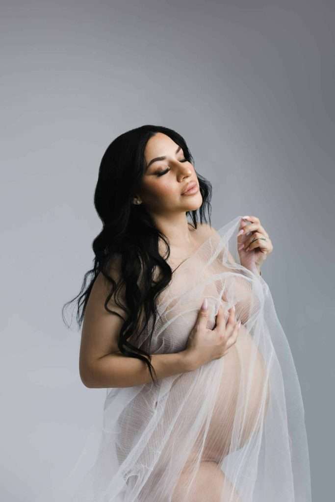 A pregnant woman with long dark hair poses draped in sheer white fabric against a light gray background.